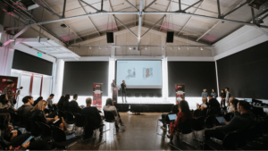 Two speakers stand on-stage in front of a small crowd at a conference event